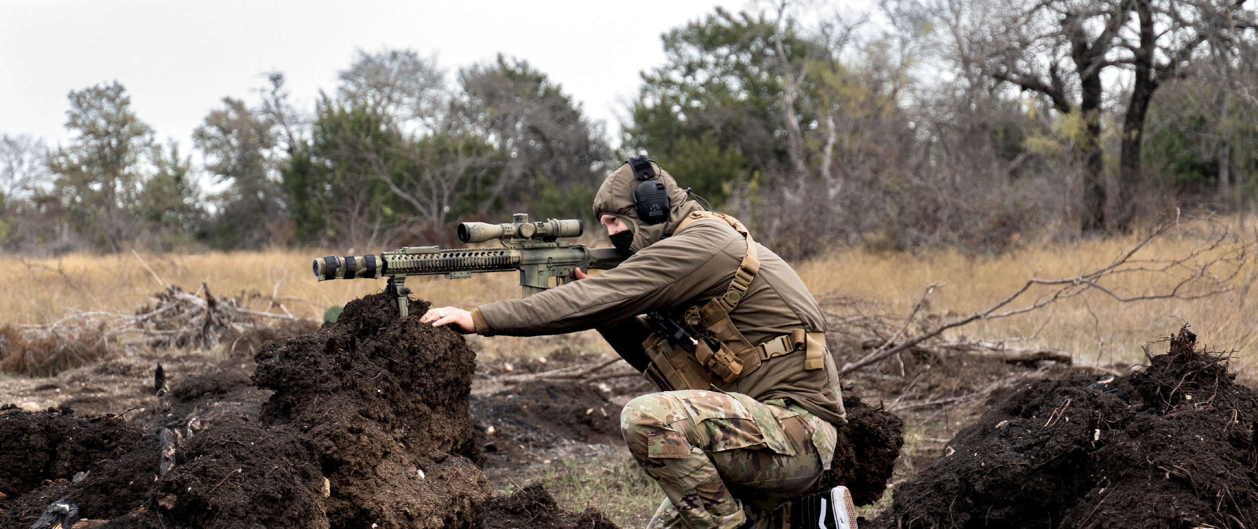 sniper in a crouched position aiming his rifle while wearing the recon chest rig