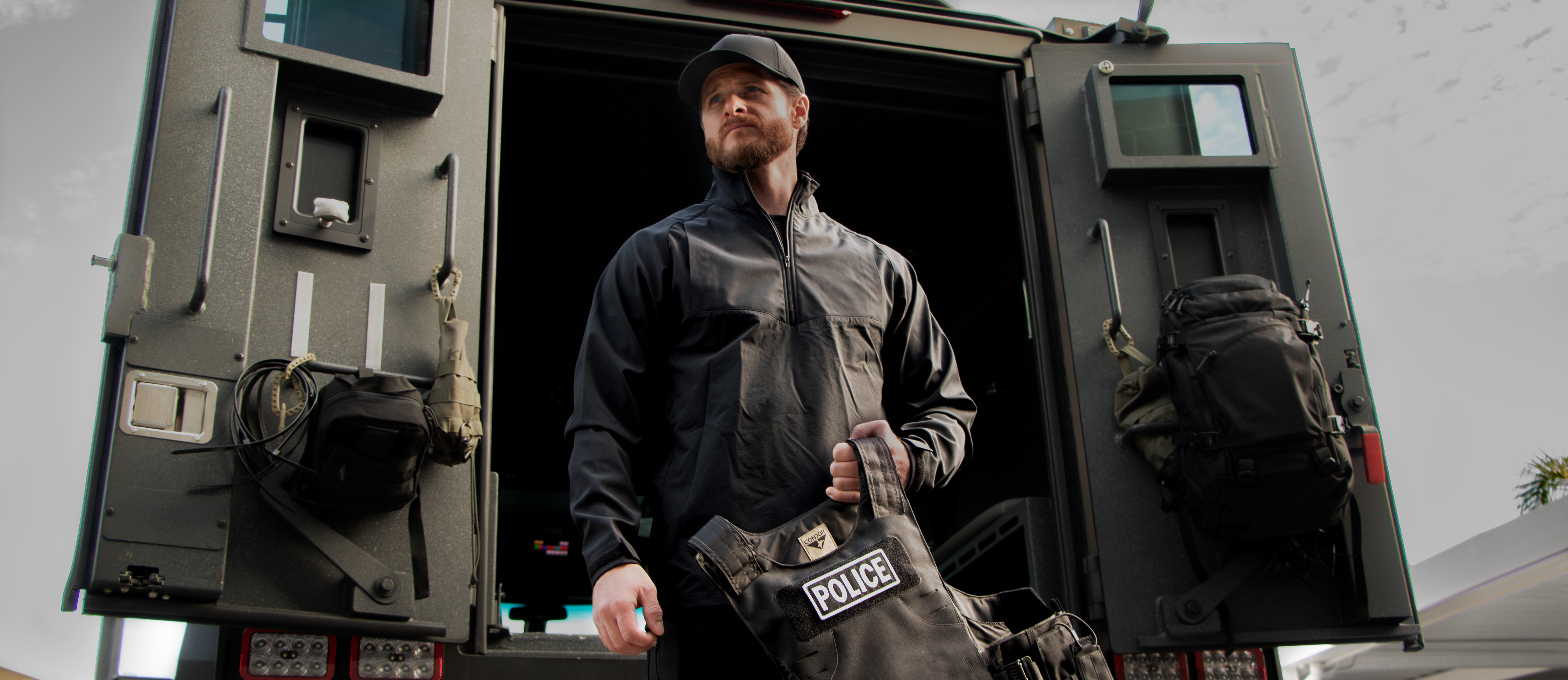 image of a police officer holding a plate carrier in front of a tectical vehicle wearing a patrol quarter zip softshell